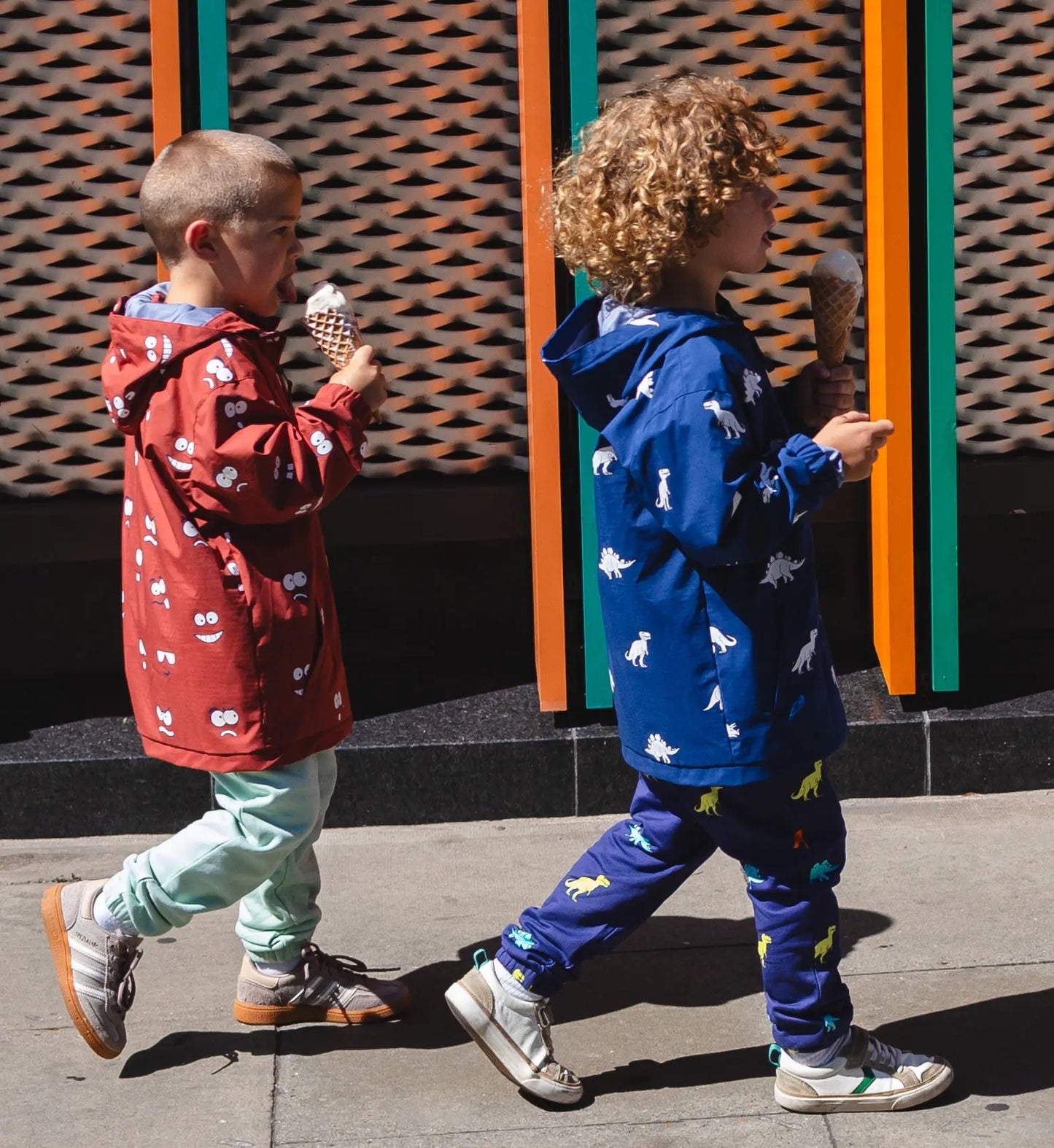 Two children in colorful outfits walking and eating ice cream against a patterned wall.