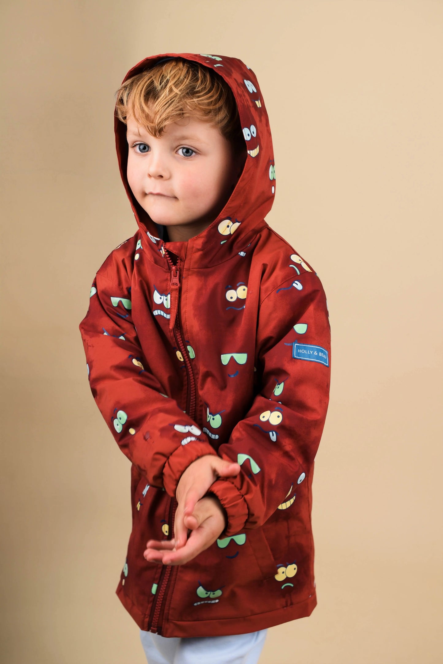 Child wearing a red hooded Eyes Colour Changing  jacket  by Holly and Beau with a eyes and faces pattern on a beige background
