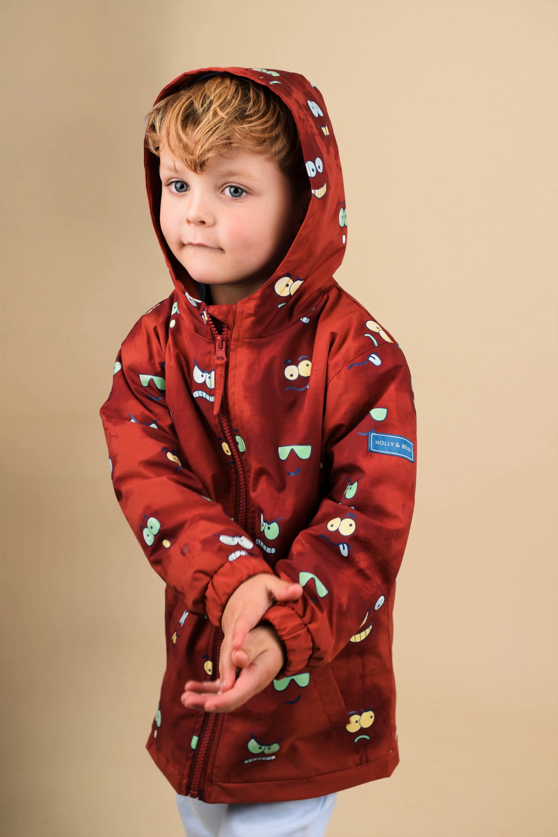 Child wearing a red hooded Eyes Colour Changing  jacket  by Holly and Beau with a eyes and faces pattern on a beige background